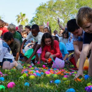 Children participating in an Easter egg hunt, enthusiastically gathering colorful plastic eggs scattered across a grassy area. The kids are crouched down with baskets and buckets, and a few adults in the background are watching and taking photos. It's a sunny day with green trees and a clear blue sky.